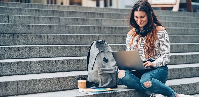 College-aged girl sitting on steps with laptop in her lap and backpack and coffee sitting beside her