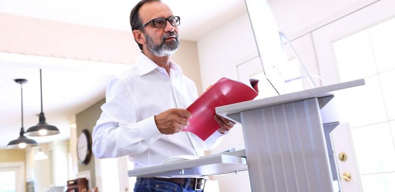 A man with glasses standing at a white standing desk in his house holding a red folder.