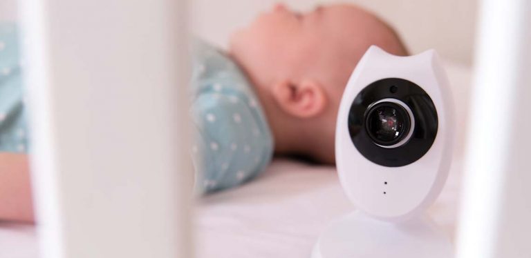 A white baby monitor between the spokes of a crib with a blurred baby in the background.
